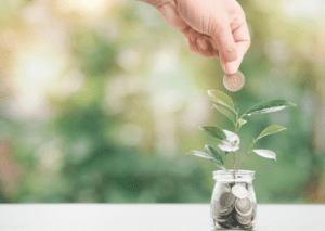 Hand-dropping a coin into a glass jar filled with coins, with a plant growing from it, symbolizing investment and growth.