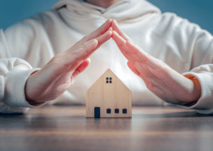 A person forming a roof shape with hands over a small wooden house model, symbolizing home safety and assurance coverage.