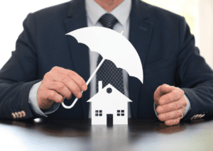 A man in a suit holding a paper umbrella over a paper house model, symbolizing home assurance and protection coverage.