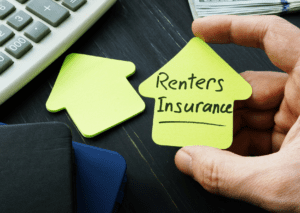 Hand holding a house-shaped sticky note with "Renters Insurance" written on it, next to a notebook and money on a desk.