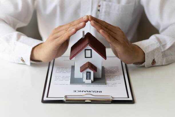 Hands protecting miniature house model on insurance document, representing Home Insurance Greeley CO coverage and protection.