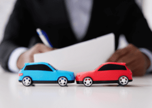 Model cars in a collision pose, symbolizing auto insurance coverage, with a person reviewing documents in the background.