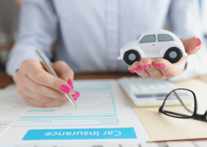 A person filling out a car insurance form while holding a toy car symbolizes auto insurance coverage.