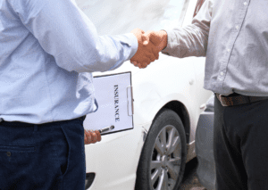 Two people shaking hands beside a car with a document in his hand, symbolizing agreement on auto coverage.